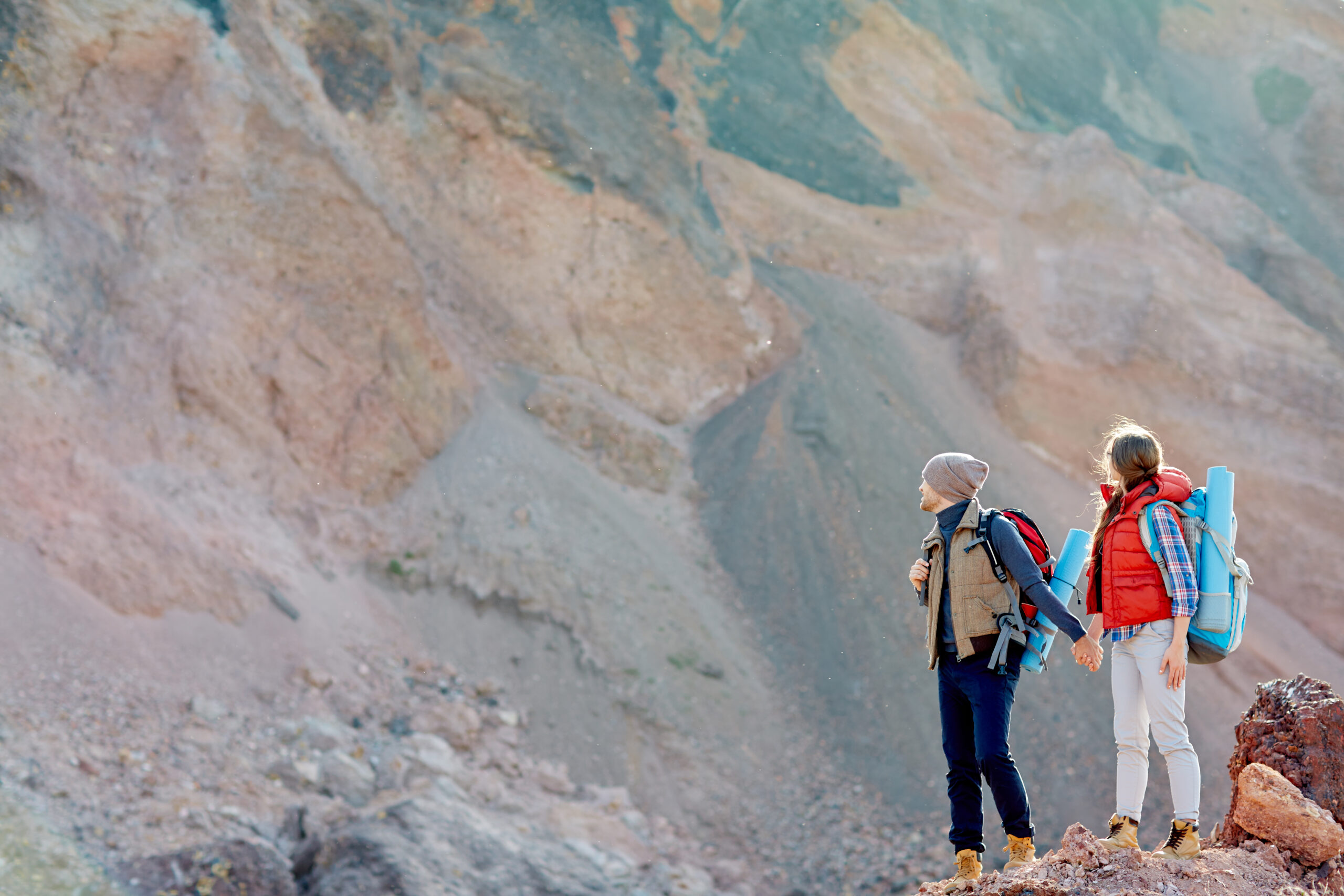 Wide shot copy space image of small figures of people in hiking gear with backpacks standing against background of enormous mountain holding hands, traveling together.